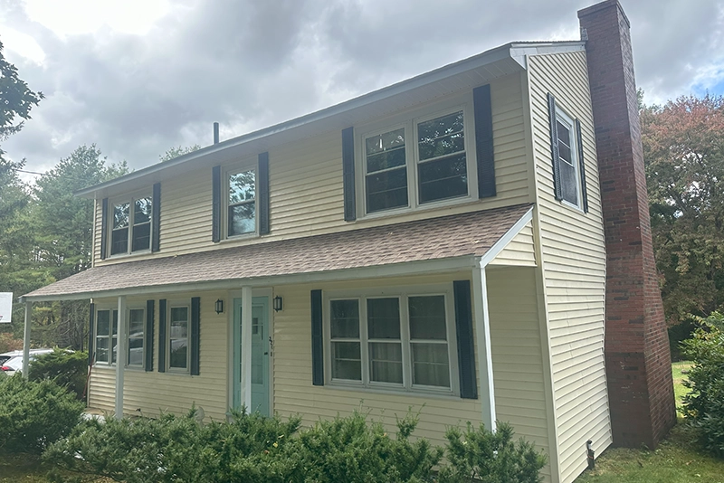 A two-story, light yellow house with black shutters and a brick chimney, surrounded by green bushes under a cloudy sky.